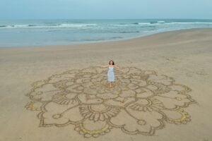 a large mandala painted on the sand on the ocean with a girl in the middle boho style.