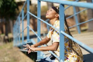 Black girl with braids sitting on a bridge enjoying the sun on her face with her eyes closed