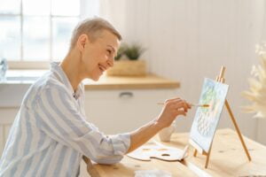 Middle aged creative woman painting picture at home while sitting at wooden table in kitchen